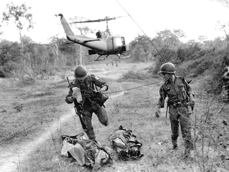 U.S. Air Force Lt. Buck Rennick, left, of Wichita, Kan., tethered to his radio man by a coiled radio line, jumps over the bodies of two South Vietnamese paratroopers while both take cover from sniper fire near Binh Gia, March 1, 1965. A helicopter used in the engagement takes off, also under fire. (AP Photo)